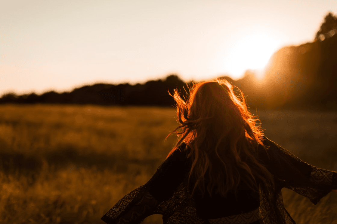 A woman with flowing red hair gracefully strolls through a sunlit field, capturing the beauty of the sunset.