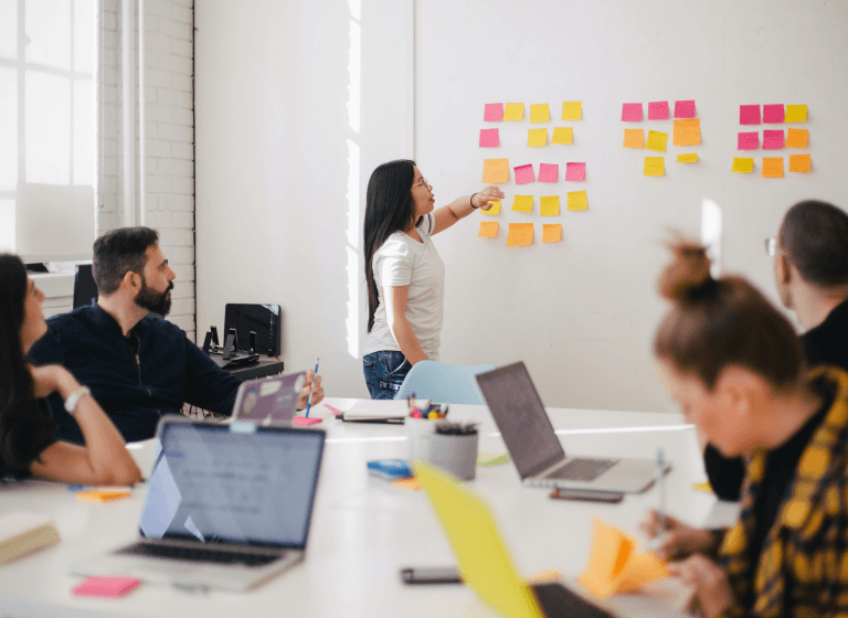 Office environment with individuals engaged in teamwork, utilizing sticky notes on the wall. Embracing critical feedback to create the finest product.