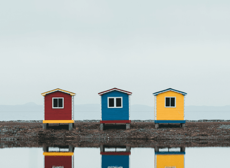 The reflection of three vibrant beach huts in water, symbolizing the Idea Meritocracy philosophy that disregards egos, labels, and titles.